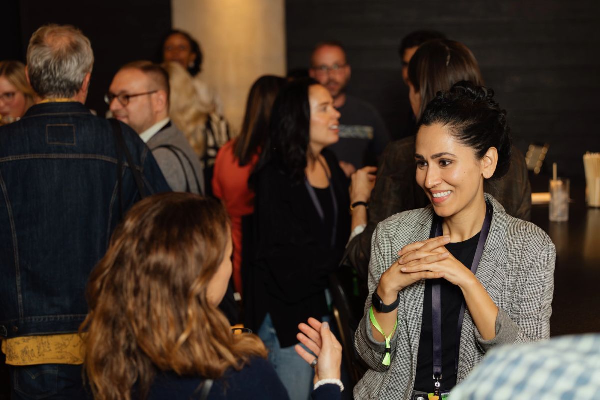 Professional woman in gray blazer speaking with seated attendee at networking event with other professionals.