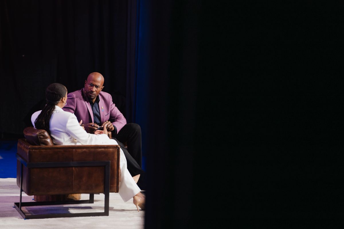Two speakers in conversation on stage, one in white outfit and one in purple blazer, seated on brown leather.