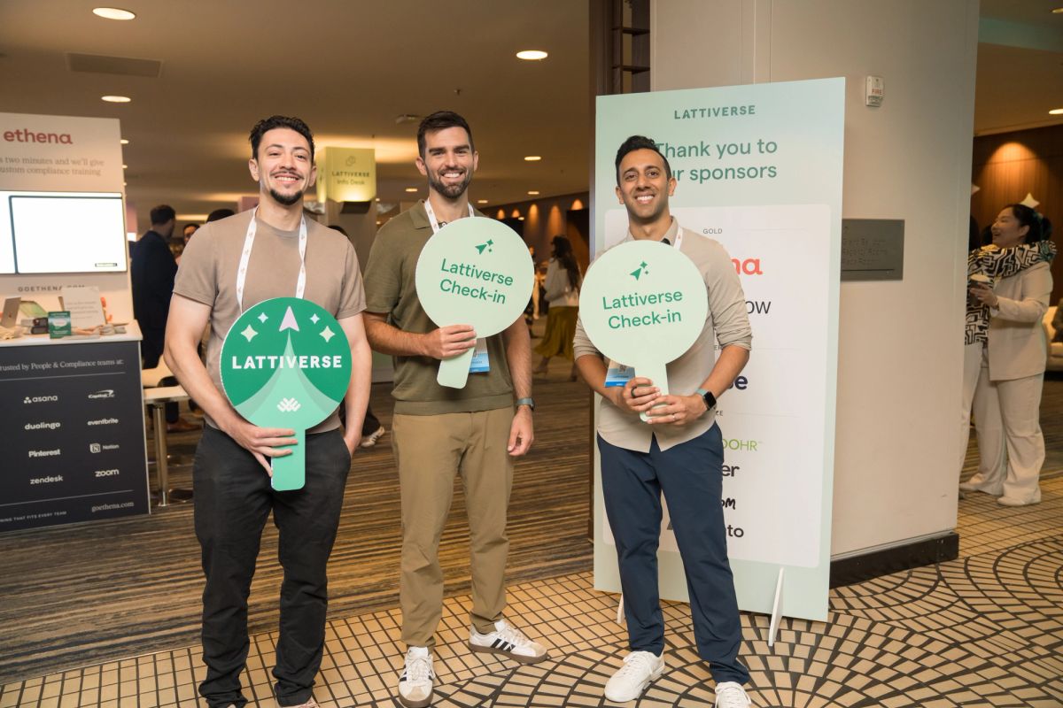 Three attendees smile holding Lattiverse signs at a conference check-in area.