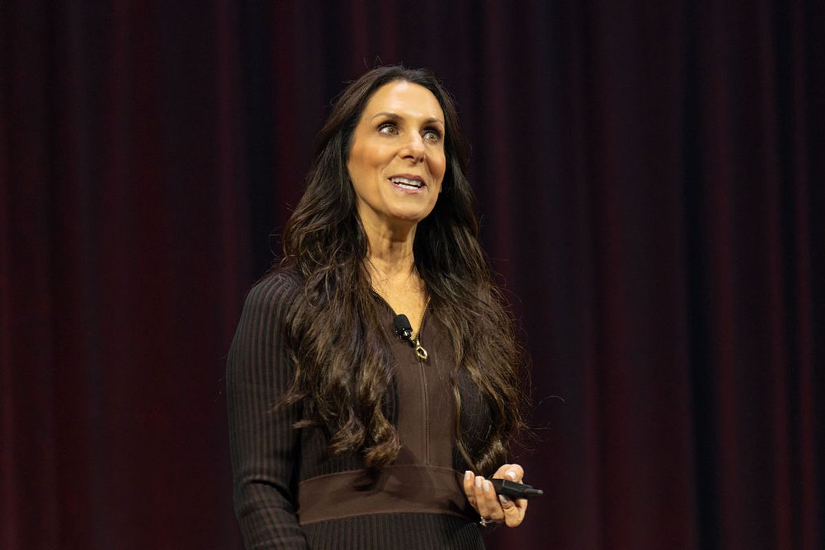 Woman with long dark hair speaking on stage wearing black outfit against dark red curtain backdrop