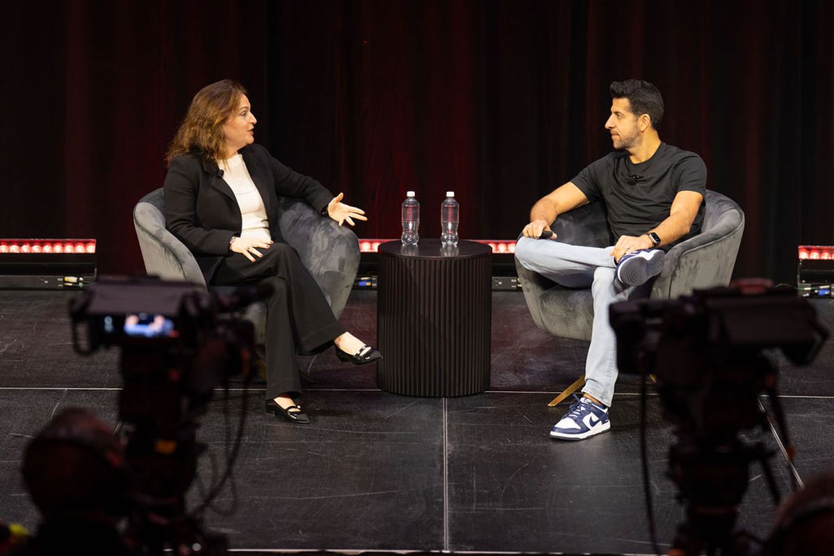 Two speakers in conversation on stage, seated in gray chairs with water bottles between them, cameras filming.
