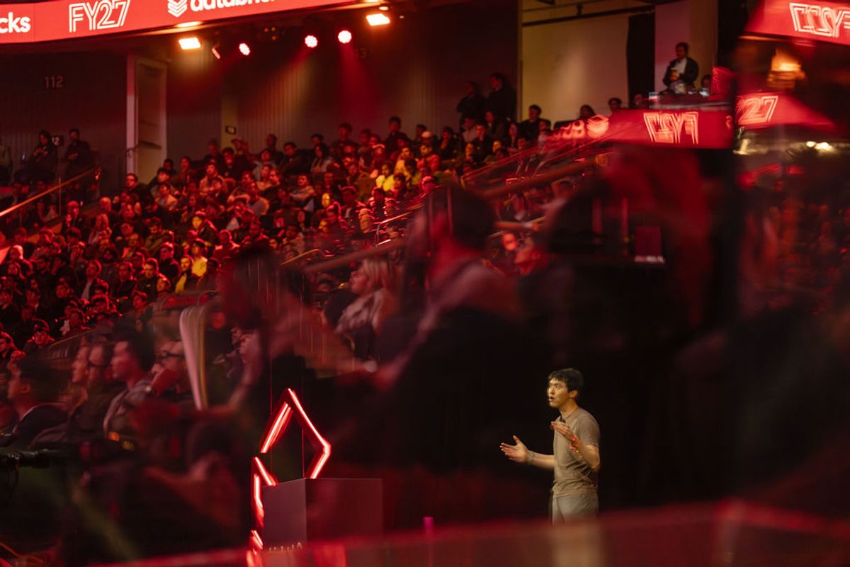 Speaker presenting on stage with hand gestures, wearing brown t-shirt against dark backdrop with red accent