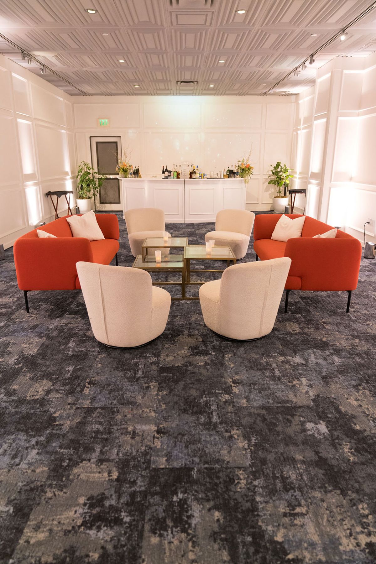 Modern lounge area with orange and beige chairs arranged around glass coffee table, white bar in background