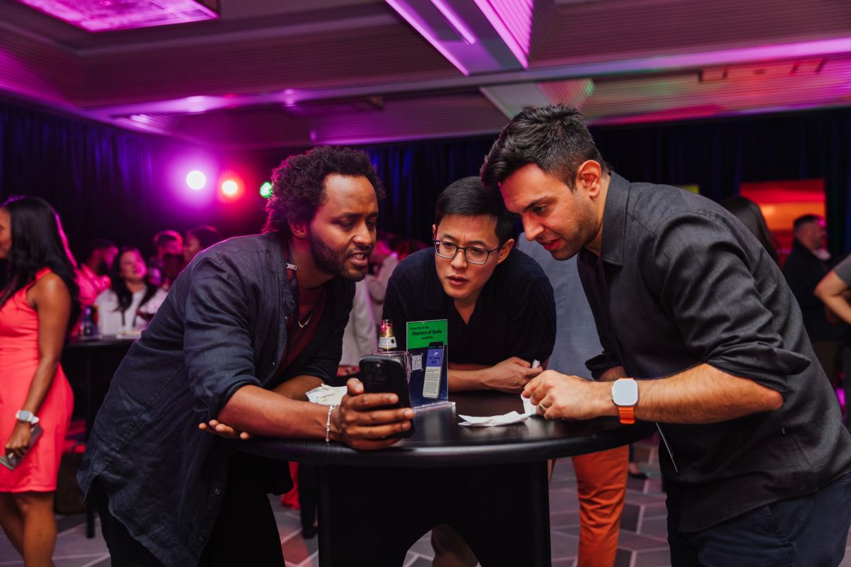 Three men gather around a table looking at a phone in a dimly lit venue with purple lighting and other.