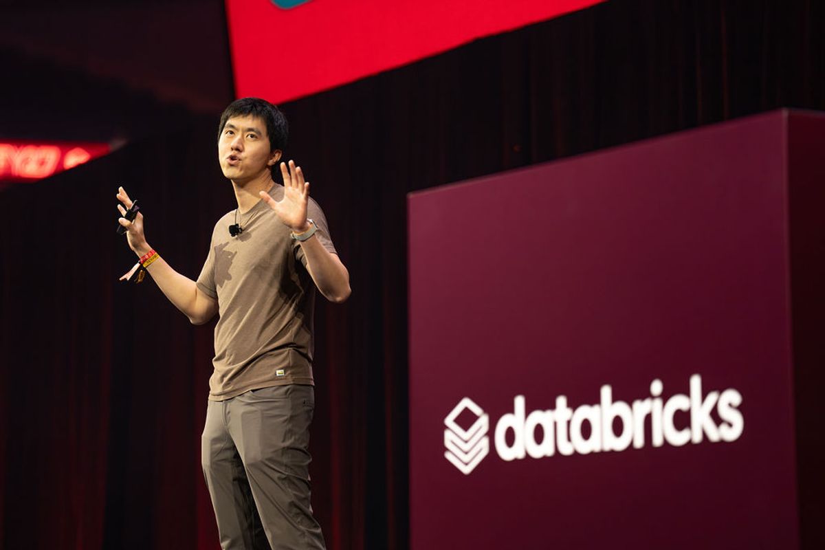 Speaker presenting on stage with microphone at Databricks conference, gesturing with hand raised