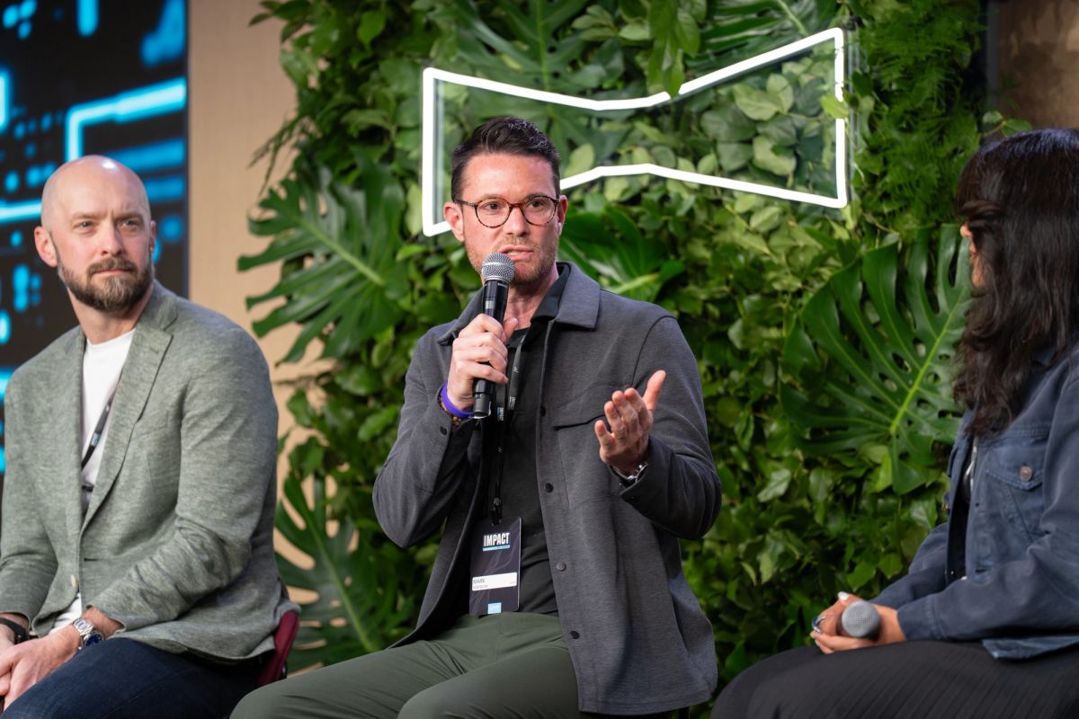 Three men on stage at panel discussion, center speaker holding microphone and gesturing, with green plant.