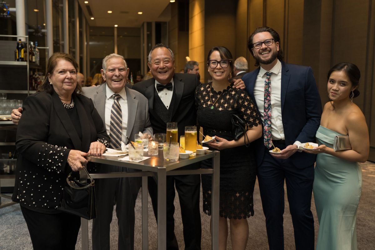 Six formally dressed people pose together at a cocktail table during an upscale evening event.