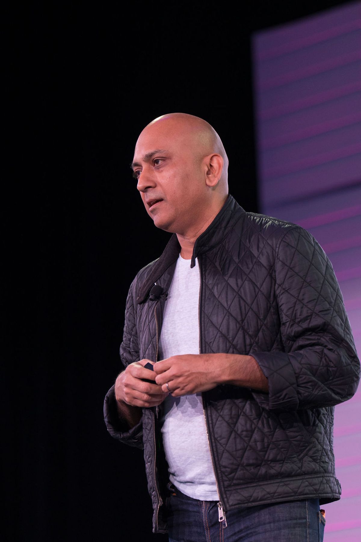 Bald speaker in black quilted jacket and white shirt presenting on stage with purple backdrop