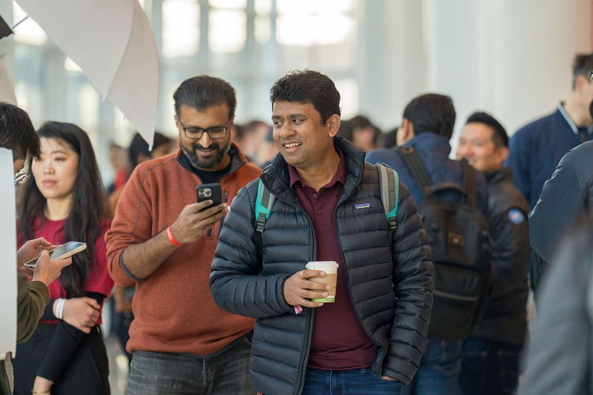 Two men walking and smiling at a conference, one holding a phone and the other carrying coffee, with other.