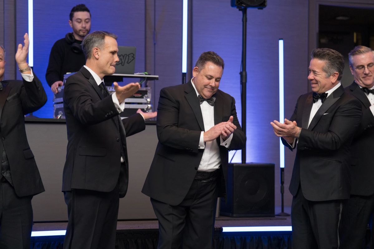 Men in tuxedos applauding and celebrating on stage at a formal gala event with blue lighting