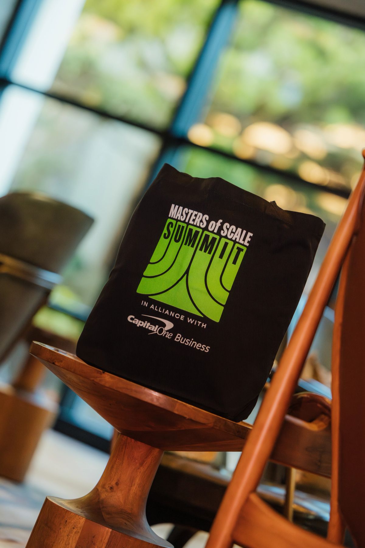 Black folder with "Masters of Scale Summit" logo sits on wooden chair at business conference venue