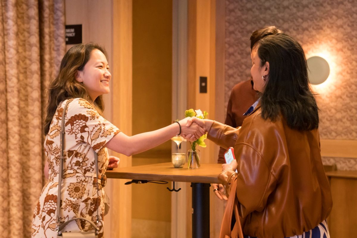 Two women shake hands at a small table during what appears to be a panel discussion or networking event.