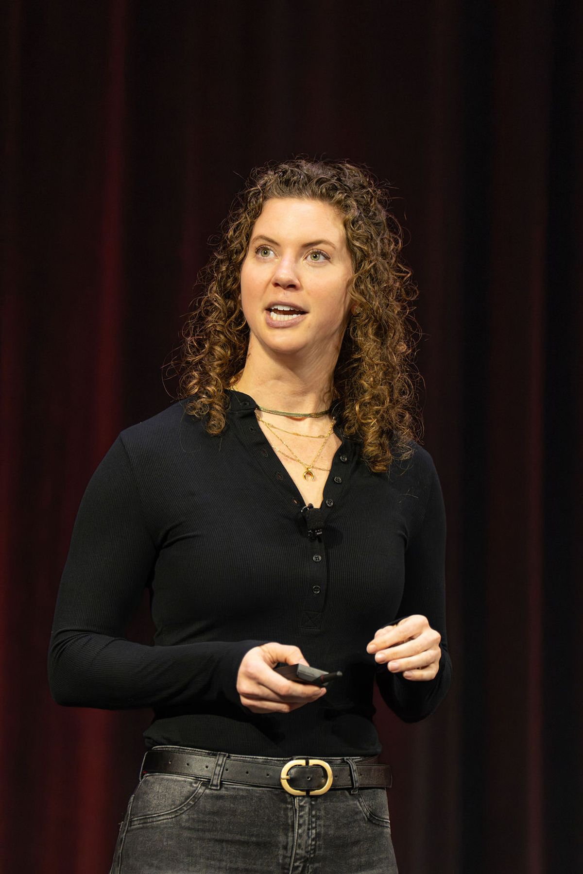 Speaker with curly hair presenting on stage, holding remote, wearing black top and jeans with belt