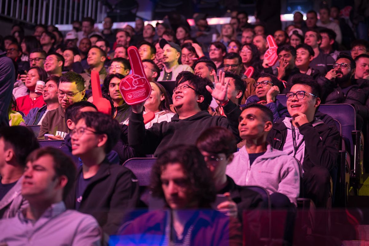 Audience members at a conference applauding and holding red foam fingers, seated in tiered seating under.