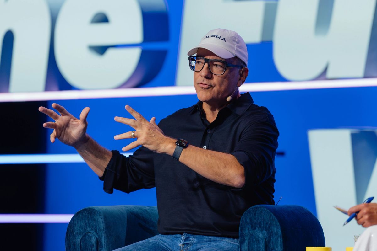 Speaker in baseball cap and glasses gestures with both hands while seated on stage at conference