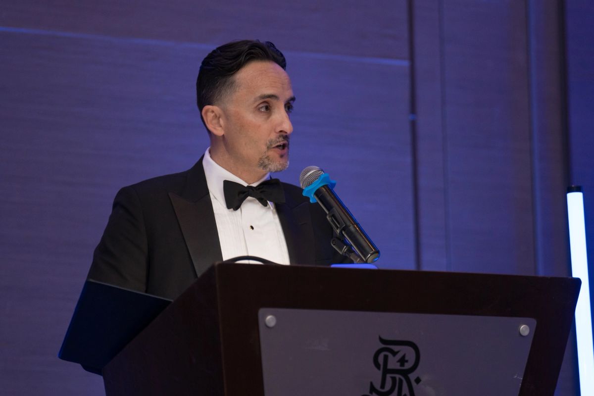 Man in black tuxedo speaking at podium with microphone during formal event with purple lighting