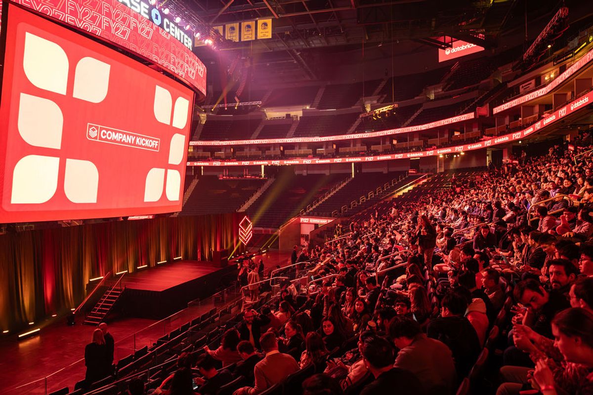 Large crowd of attendees in arena seating watching presentation on big screen with red stage lighting
