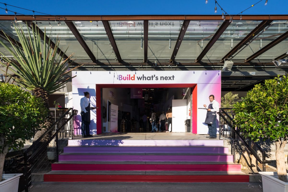 Event stage with red and purple steps leading to white booth displaying "Build what's next" banner