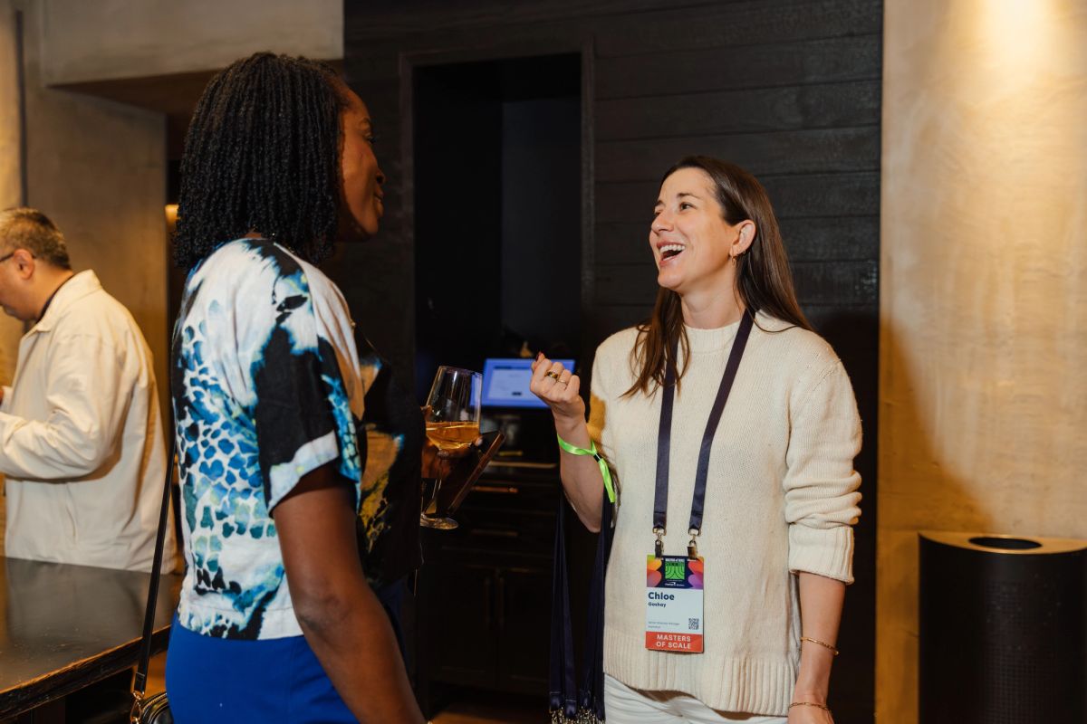 Two women networking at a conference, one holding a drink and name badge, both smiling in conversation.
