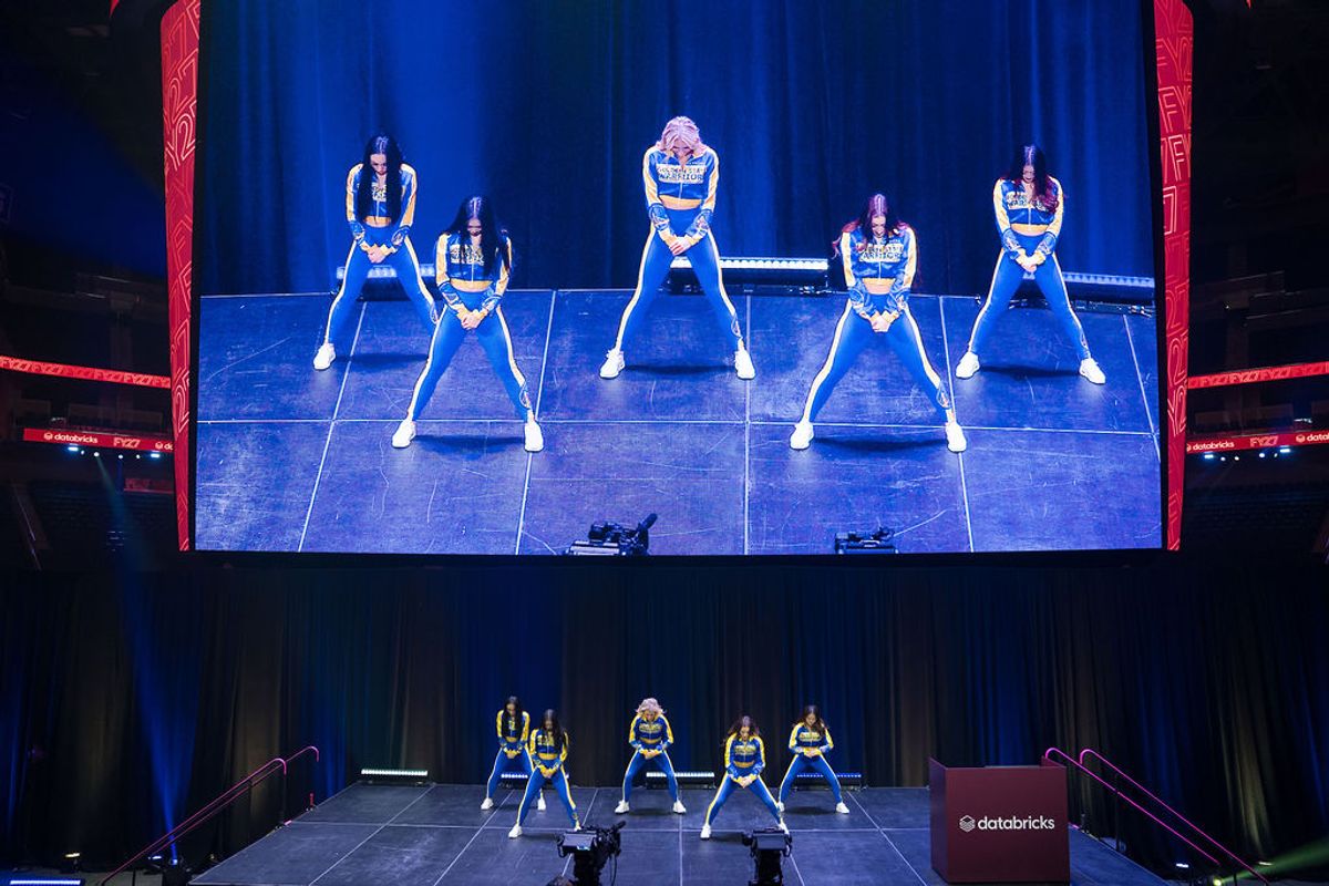 Five cheerleaders perform synchronized routine on stage with large video screen displaying their performance.