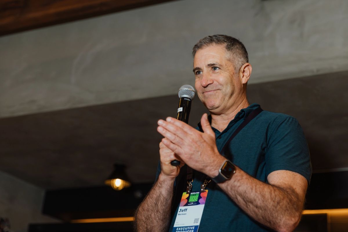 Executive in teal shirt speaking into microphone while gesturing at indoor conference venue