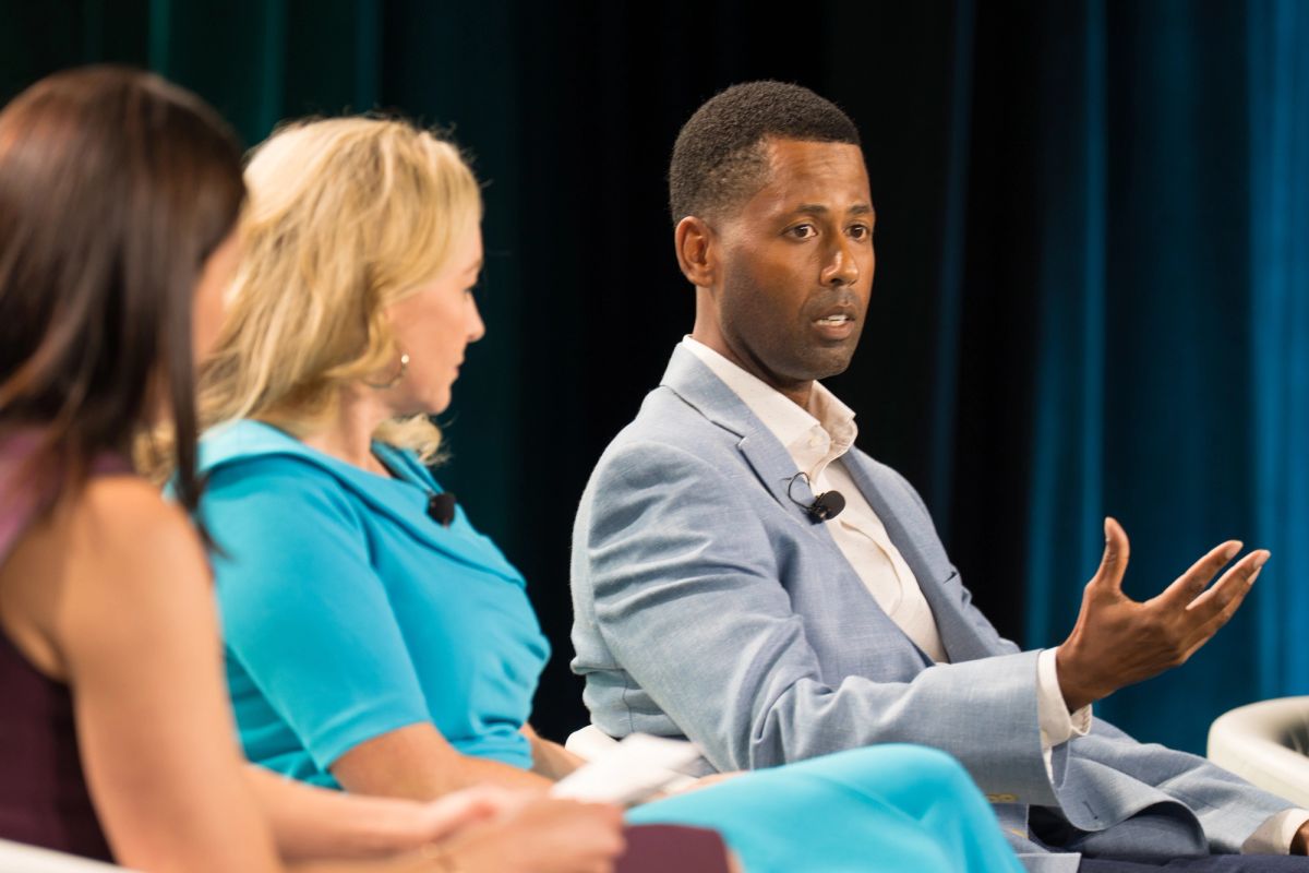Three panelists seated on stage, one man speaking and gesturing during a conference panel discussion.