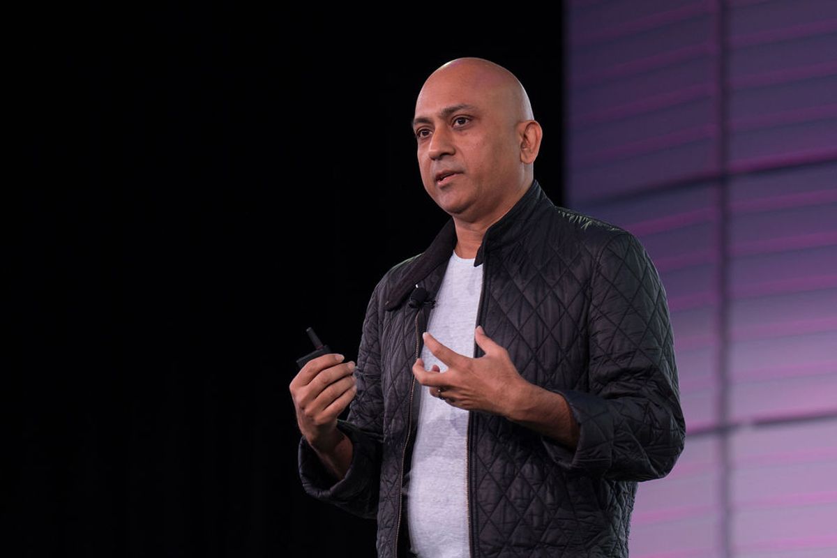 Bald speaker in black quilted jacket holding microphone gestures while presenting on stage with purple.