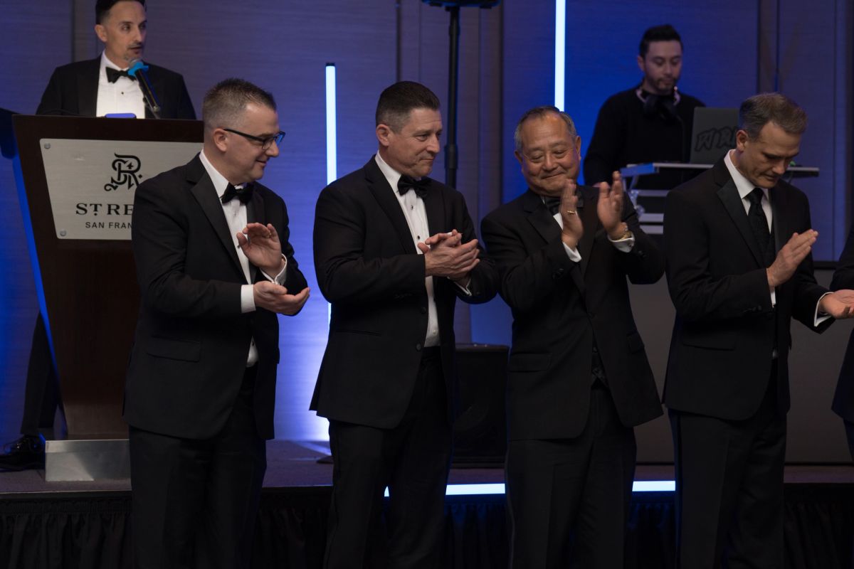 Four men in tuxedos applauding at formal gala event with podium and blue lighting at St. Regis venue
