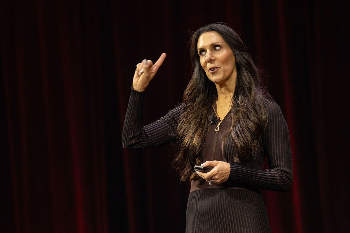 Speaker with long dark hair gesturing while presenting on stage with dark curtained backdrop