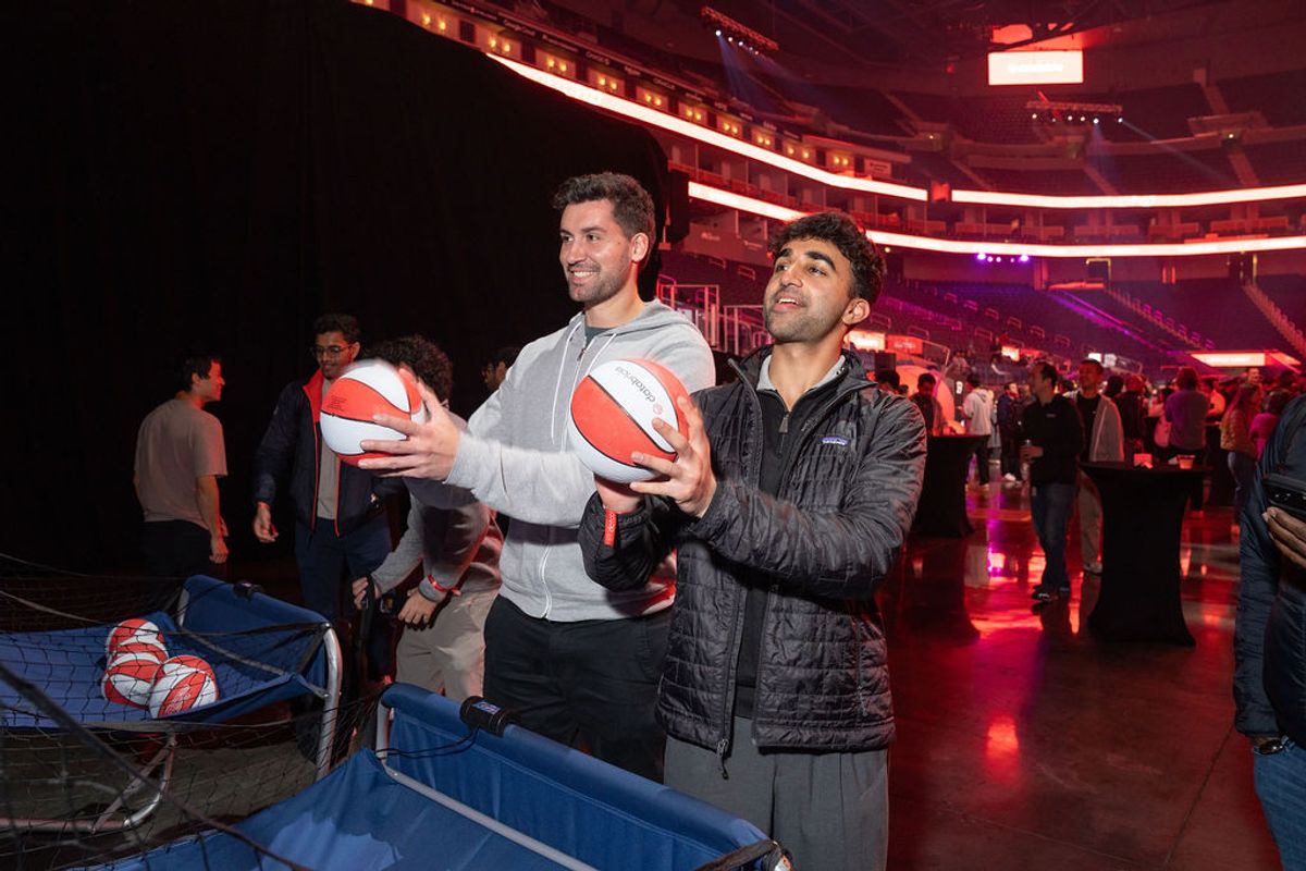 Two men hold basketballs while standing near blue storage bins in a large arena with red lighting and crowds