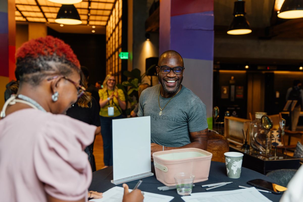 Two people at a conference table, one person signing documents while a smiling man in glasses watches from.