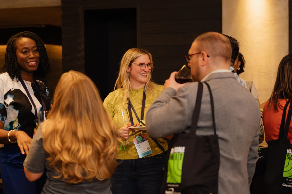 Conference attendees networking and conversing in a modern venue with columns and warm lighting.