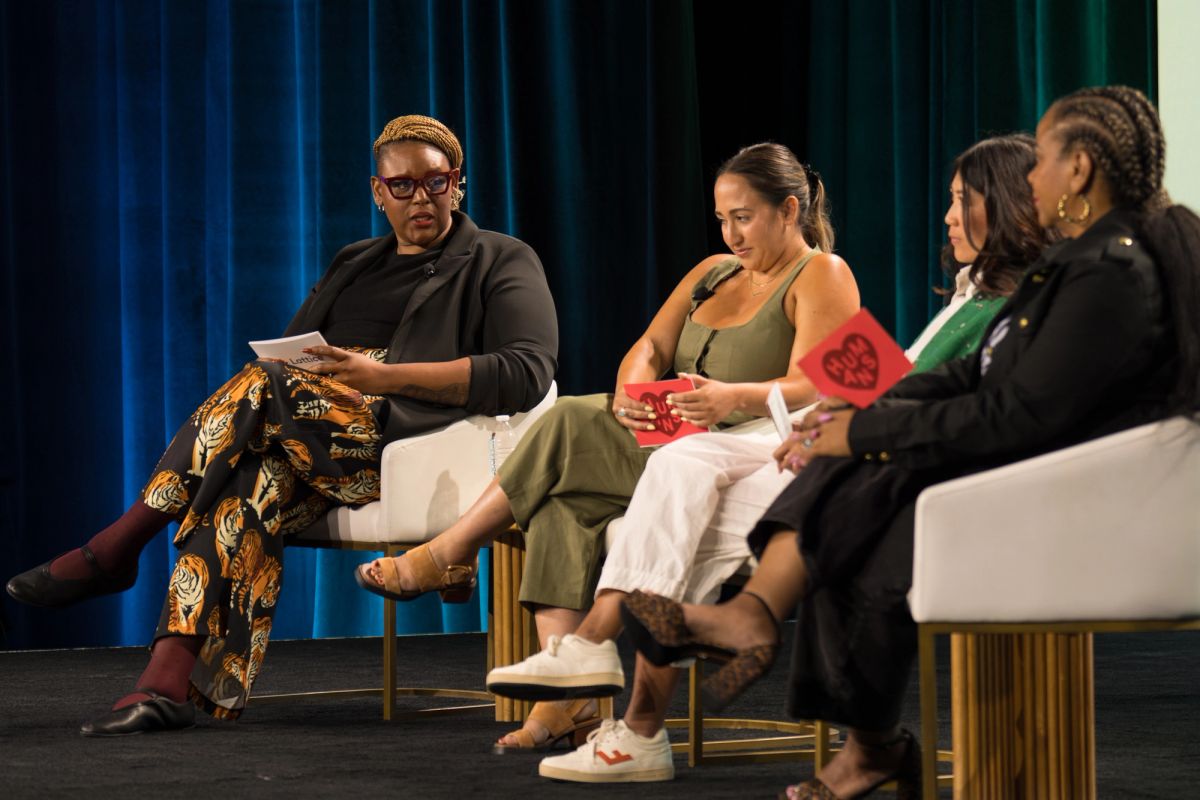 Four women seated on stage in discussion panel, teal curtain backdrop, holding note cards.