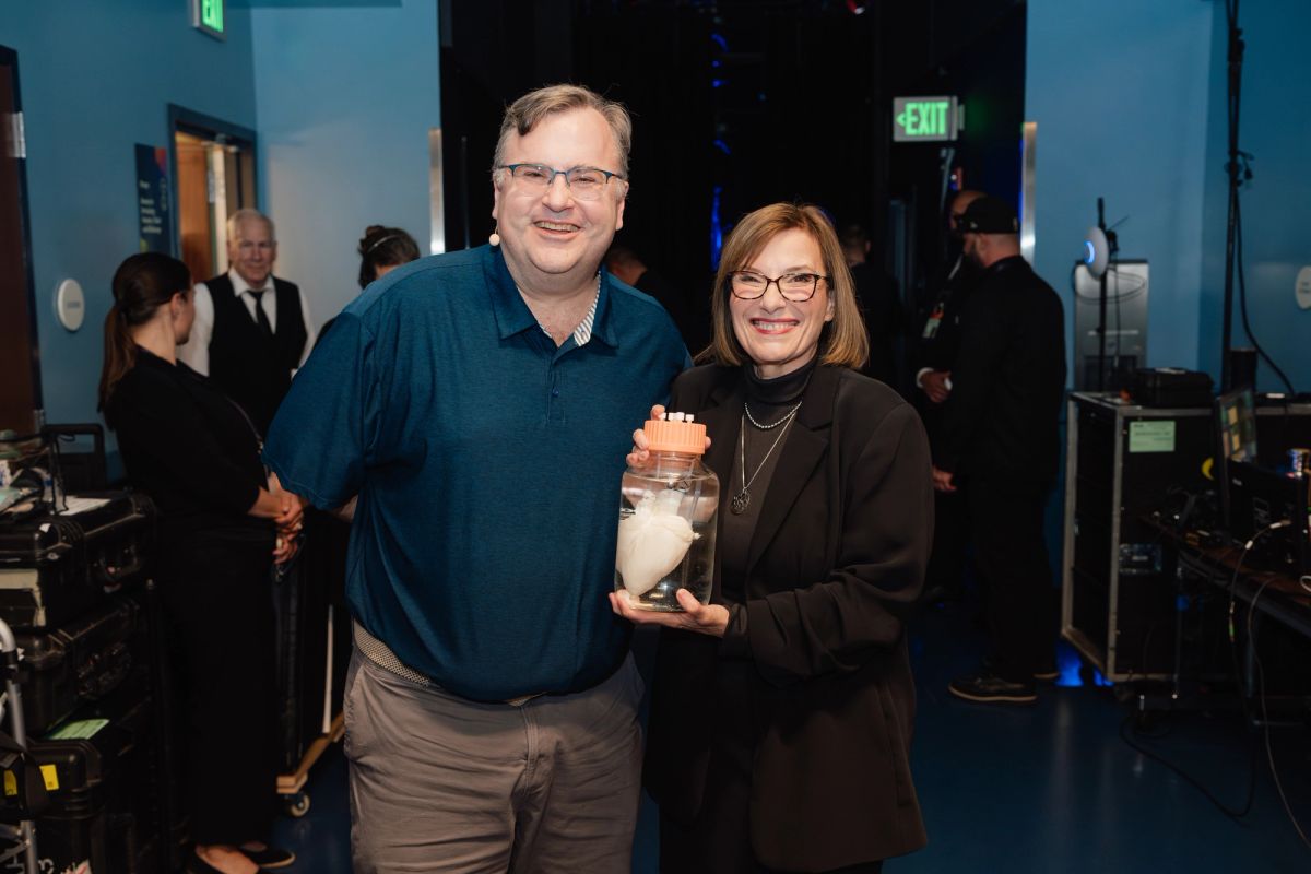 Two people pose together at an indoor event, with the woman holding a decorative jar or container.