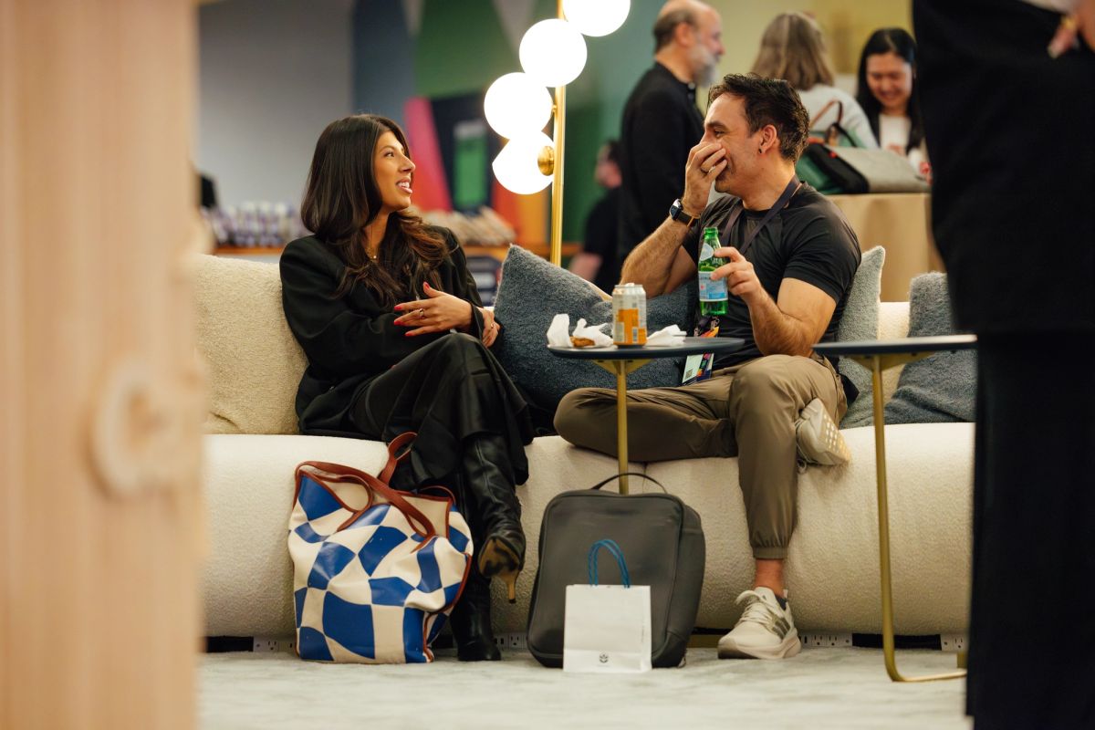 Two conference attendees sit on white couches having a conversation in a modern lounge area with luggage.
