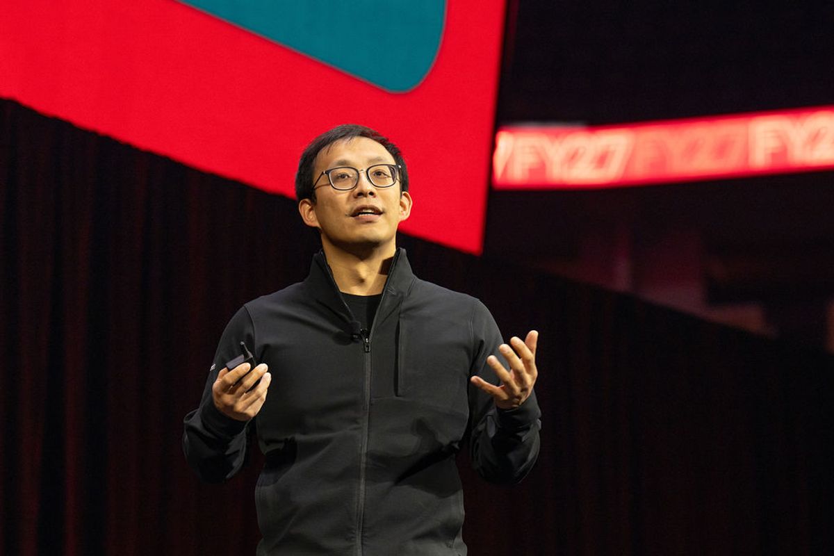 Speaker in black jacket presenting on stage with hands raised, red and teal geometric backdrop behind him