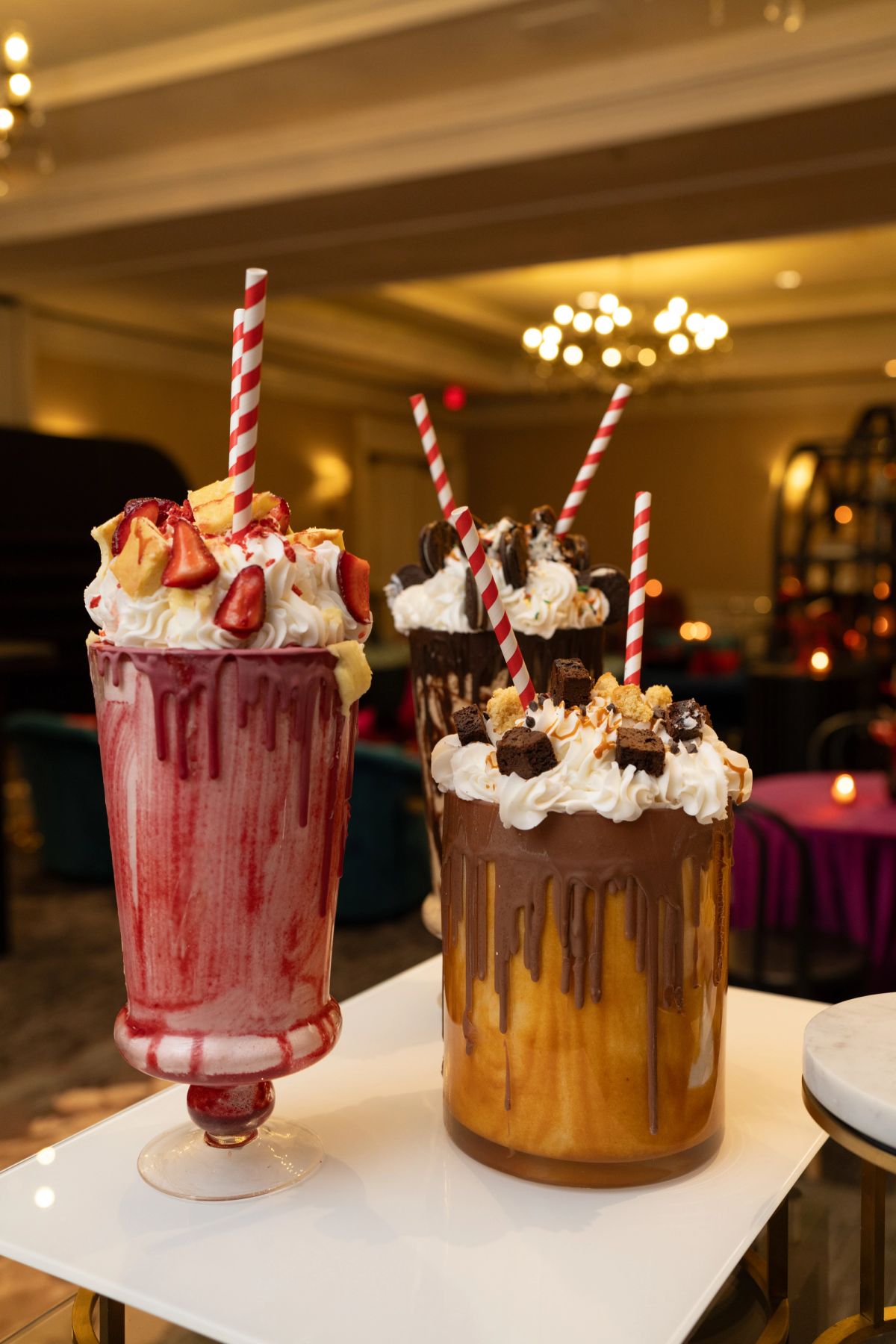 Two elaborate milkshakes with whipped cream, toppings, and striped straws on a white table in an upscale.
