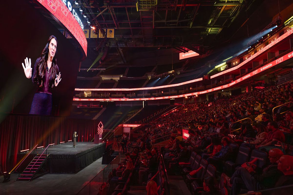 Speaker presenting on stage to large arena audience with red lighting and large screen display
