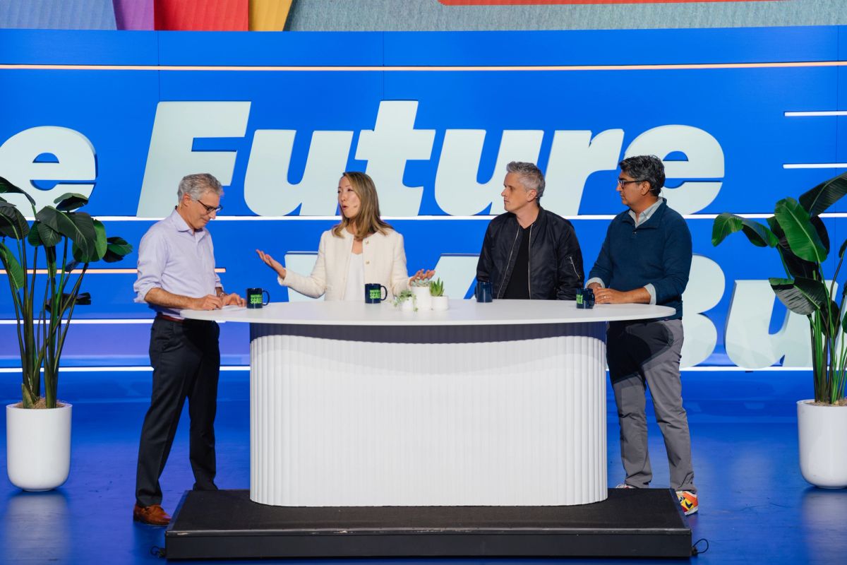 Four people on stage at conference, three standing behind white curved table, one presenting, blue backdrop.
