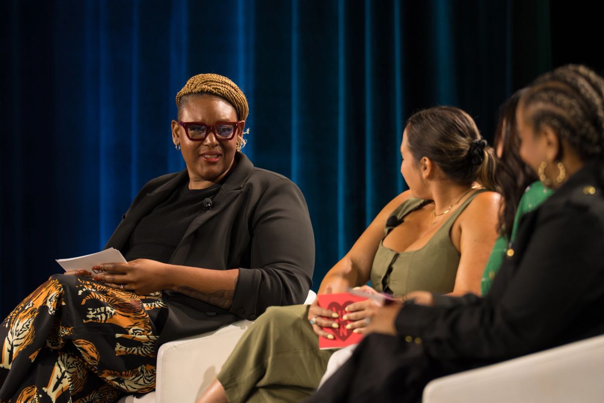 Three women seated on stage in discussion panel, blue curtain backdrop.