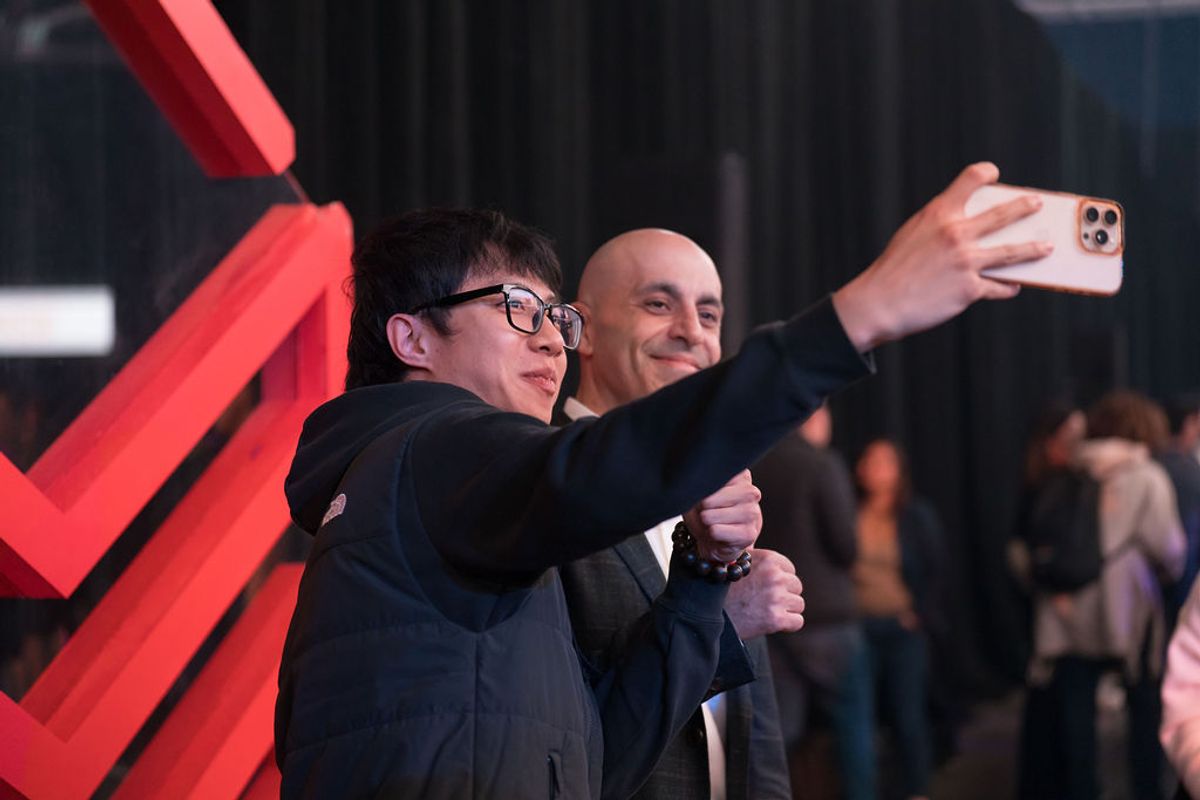 Two men in suits taking a selfie with a smartphone at a business conference, with red signage visible in the.