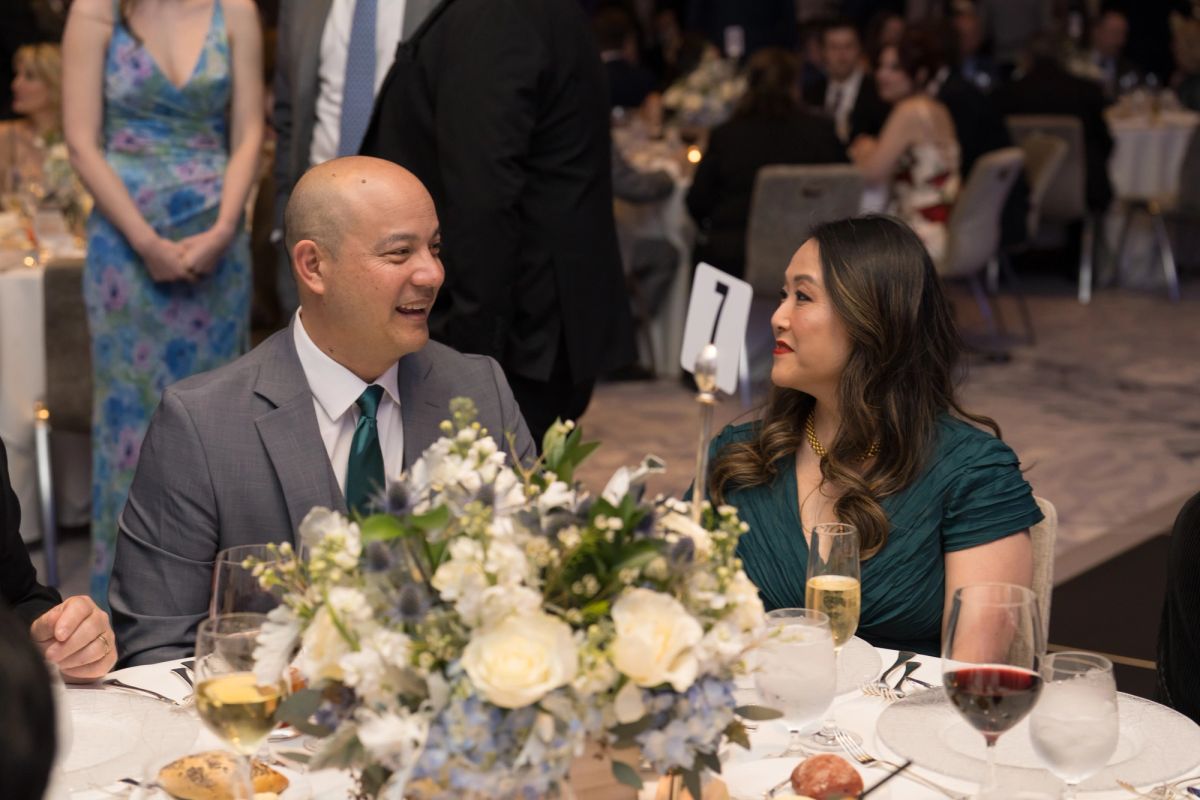Two people in formal attire sitting at elegantly set dinner table with white floral centerpiece and wine.