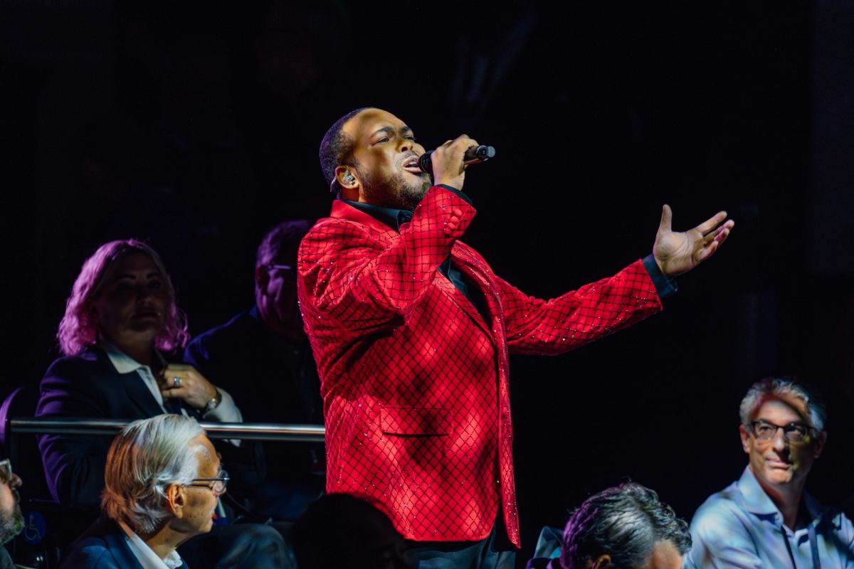 Singer in red jacket performing on stage with microphone while audience members watch from theater seating
