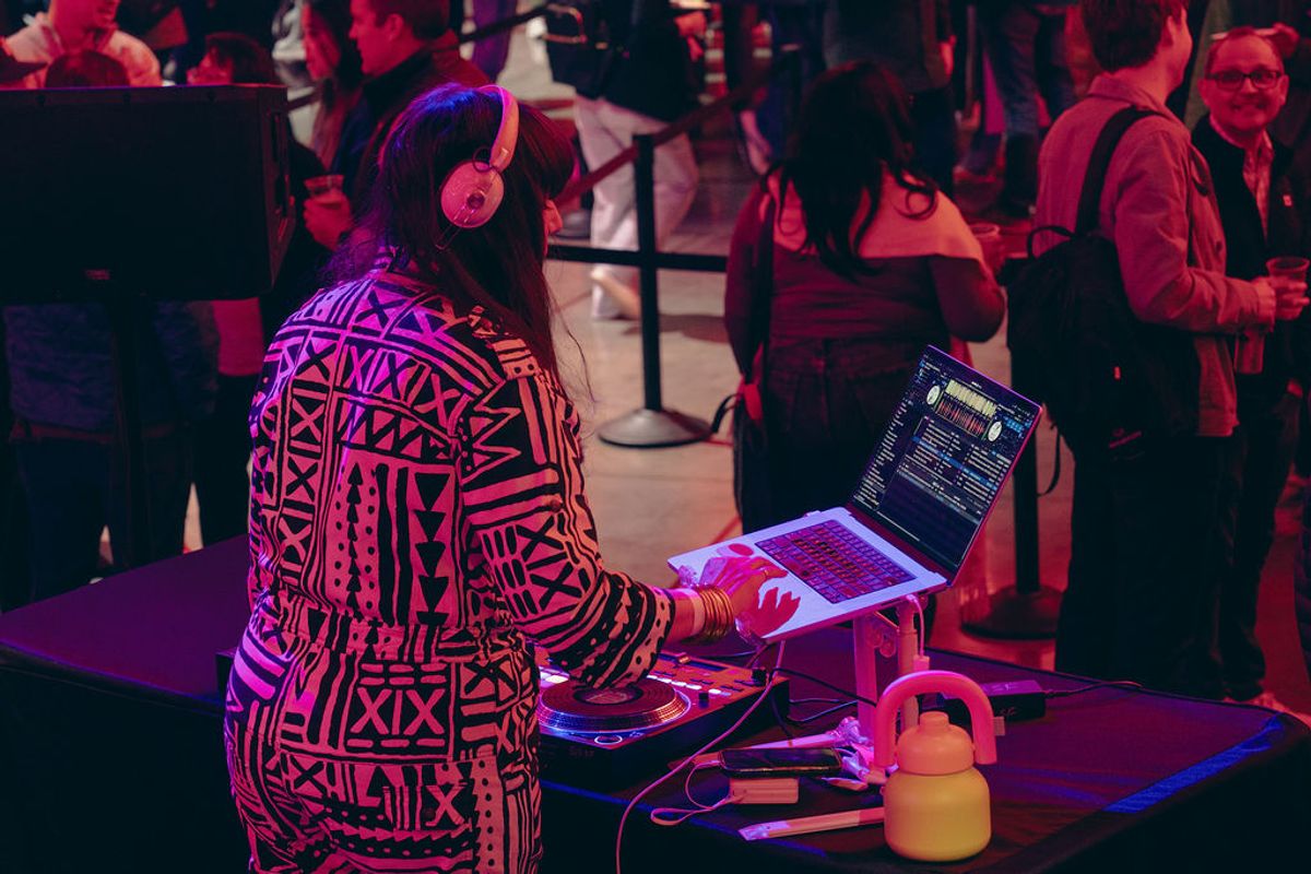 DJ with headphones and patterned jacket performs at laptop setup while conference attendees mingle in.