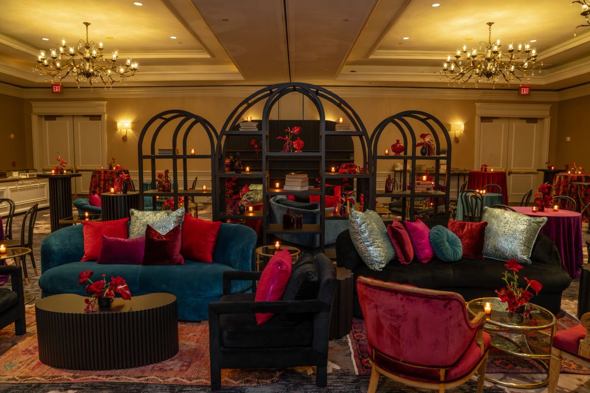 Elegant hotel lobby lounge with teal and red velvet furniture, ornate bookshelves, chandeliers, and warm.