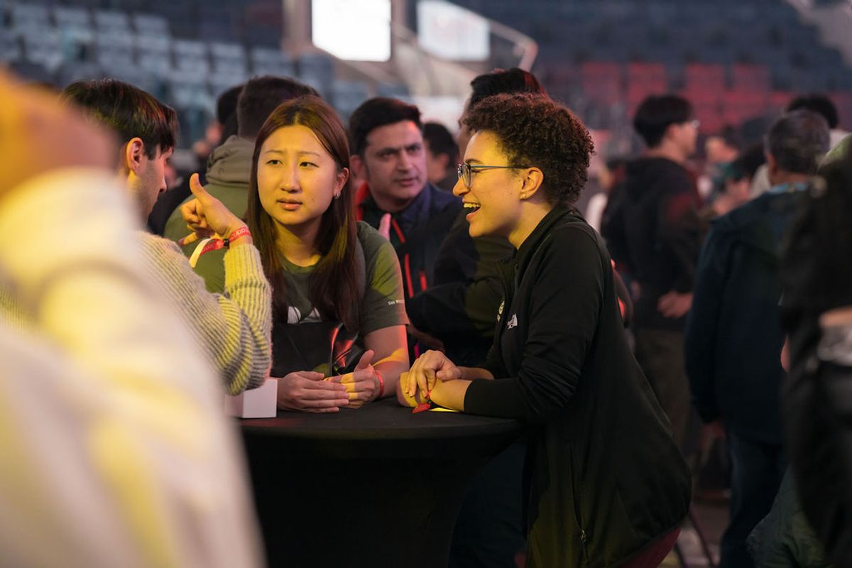 Two attendees sit at a table networking at a conference, with other participants mingling in the background