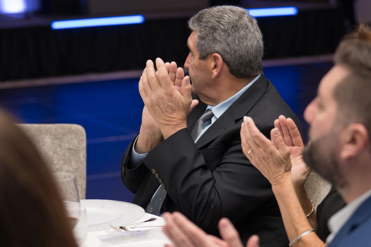 Man in dark suit applauding while seated at formal event, with other attendees clapping in background