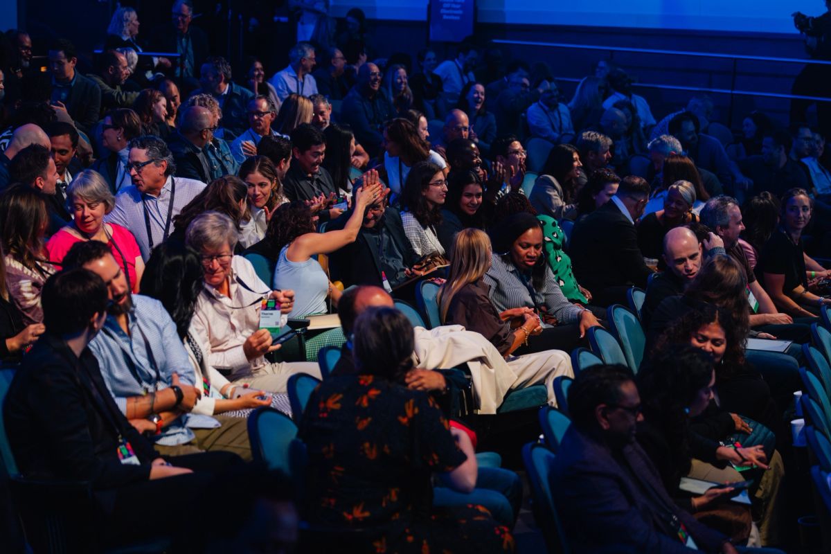 Audience members seated in blue theater chairs during a conference presentation in dim lighting