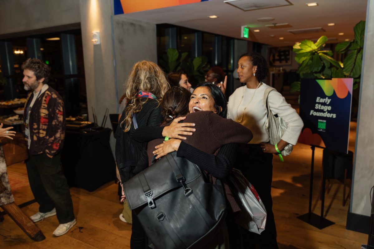 Two women embrace warmly at a networking event while other attendees mingle in a modern conference space.