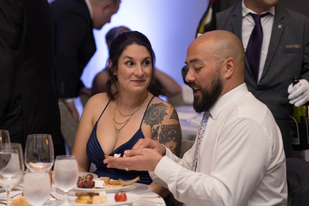 Woman in navy dress and man in white shirt conversing at formal dinner table with wine glasses and appetizers